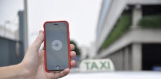 uma mão segurando um celular com o símbolo da uber e atrás um carro com a placa de taxi sobre o teto