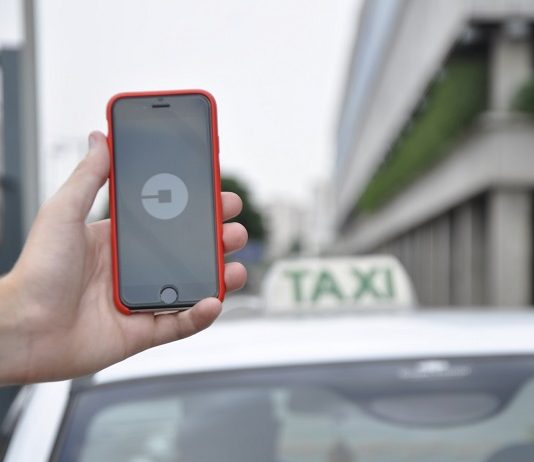 uma mão segurando um celular com o símbolo da uber e atrás um carro com a placa de taxi sobre o teto