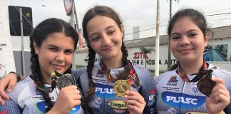 três moças com uniformes iguais de competição posam sorridentes para a câmera mostrando as medalhas penduradas no pescoço
