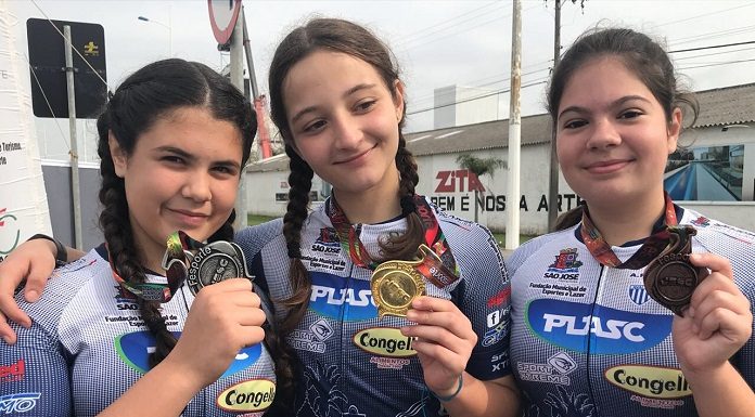 três moças com uniformes iguais de competição posam sorridentes para a câmera mostrando as medalhas penduradas no pescoço