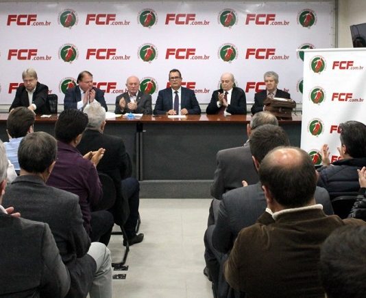 pequena sala de auditório com pessoas sentadas vistas de costas e ao fundo, na bancada, o presidente eleito ao centro de outras pessoas; todas são homens