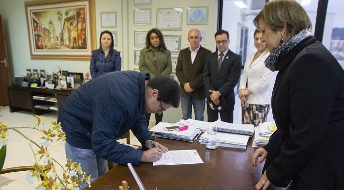 homem se abaixa para assinar um documento em uma mesa na frente da prefeita adeliana e mais algumas pessoas ao fundo em pé observam