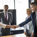 dois jovens se cumprimentam sobre uma mesa de trabalho com outras pessoas olhando em volta