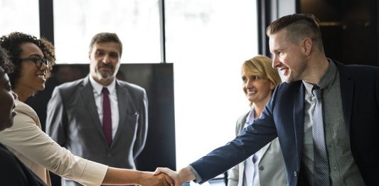 dois jovens se cumprimentam sobre uma mesa de trabalho com outras pessoas olhando em volta