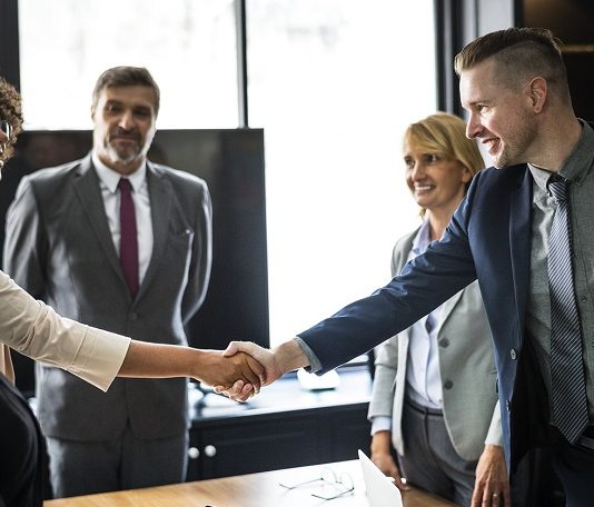 dois jovens se cumprimentam sobre uma mesa de trabalho com outras pessoas olhando em volta