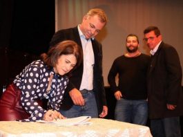 mulher assina documento em cima de uma mesa e ao lado o prefeito gean loureiro observa; há outros dois homens atrás