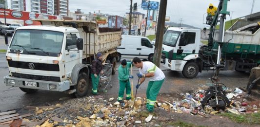 dois funcionários da comcap recolhem lixo com pás ao lado de dois caminhões da companhia