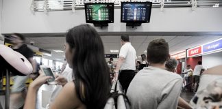 homem e mulher sentados em cada lado de bancos de espera no saguão do aeroporto; há dois monitores em uma parede no alto ao fundo