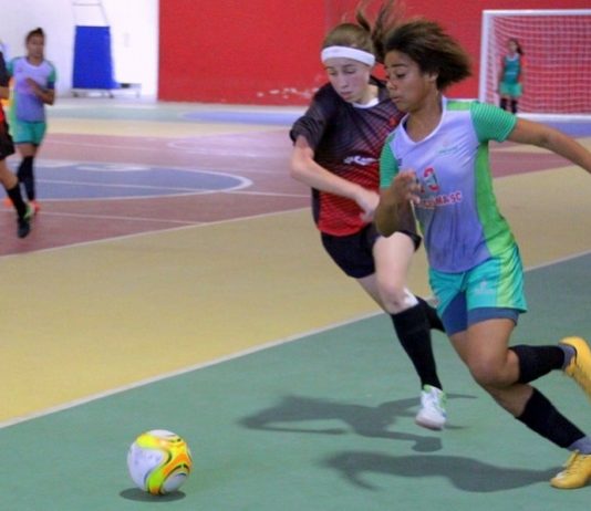 duas garotas de times diferentes correm para disputar a bola em quadra de futsal