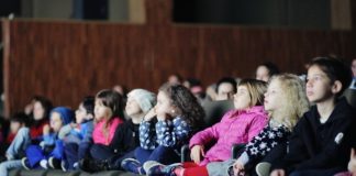 crianças em uma fileira de poltronas de cinema assistindo a um filme com os rostos iluminados pela tela; olha na mesma direção