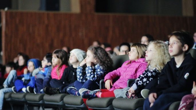 crianças em uma fileira de poltronas de cinema assistindo a um filme com os rostos iluminados pela tela; olha na mesma direção