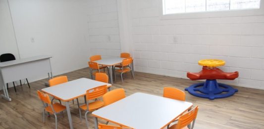 sala de aula com duas mesas infantis cercadas por cadeiras laranjas para crianças e alguns brinquedos
