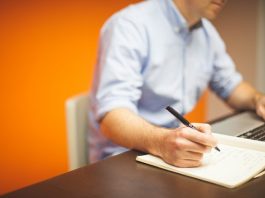 homem em frente ao notebook anota com uma caneta em um papel