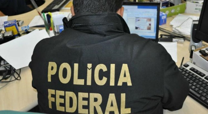 agente visto de costas sentado em uma bancada com laptop usando colete escrito polícia federal