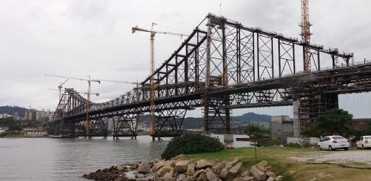 ponte hercílio luz vista da cabeceira continental em construção em dia nublado; há guindastes ao longo da estrutura