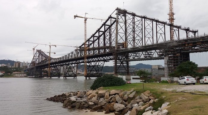 ponte hercílio luz vista da cabeceira continental em construção em dia nublado; há guindastes ao longo da estrutura