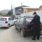 agente de trânsito visto de costas colocando um adesivo da prefeitura em um fiat uno abandonado em uma rua, com um veículo gol da prefeitura estacionado ao lado