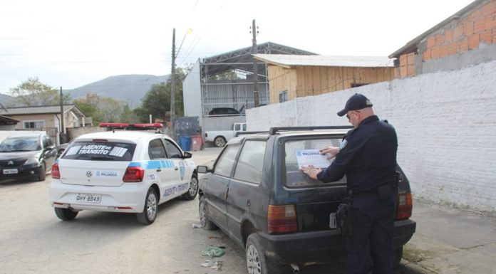 agente de trânsito visto de costas colocando um adesivo da prefeitura em um fiat uno abandonado em uma rua, com um veículo gol da prefeitura estacionado ao lado