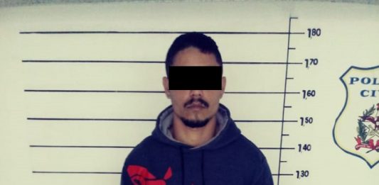 Liderança do PGC é presa ao ir à academia em Palhoça homem com o rosto censurado diante do quadro de medição de altura na delegacia posa para foto usando moletom