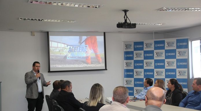 homem discursa em pé com um microfone ao lado de uma projeção de tela e à frente de uma mesa com pessoas sentadas que assitem, estão de costas para a câmera