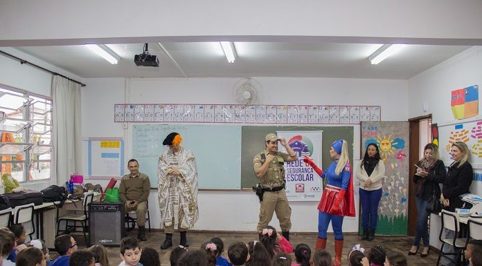 em uma sala de aula, crianças pequenas sentadas no chão assistem a apresentação de uma mulher vestida de super mulher e um policial militar de farda; ha outras professoras de pé assistindo; a maioria está sorrindo