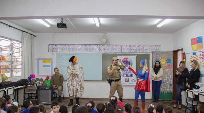 em uma sala de aula, crianças pequenas sentadas no chão assistem a apresentação de uma mulher vestida de super mulher e um policial militar de farda; ha outras professoras de pé assistindo; a maioria está sorrindo