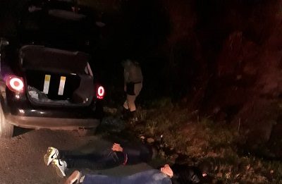 três homens deitados em estrado com as mãos algemadas para trás; há um carro e policias ao fundo; é noite