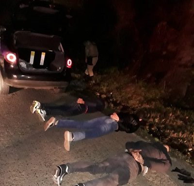 três homens deitados em estrado com as mãos algemadas para trás; há um carro e policias ao fundo; é noite