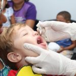 criança com os fechados e boca aberta pra cima coloca a língua pra fora ao receber gotas de vacina seguradas por mãos usando luvas; uma mulher segura a criança