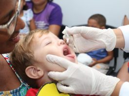 Últimos dias da campanha de multivacinação criança com os fechados e boca aberta pra cima coloca a língua pra fora ao receber gotas de vacina seguradas por mãos usando luvas; uma mulher segura a criança