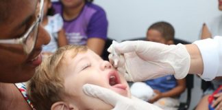 criança com os fechados e boca aberta pra cima coloca a língua pra fora ao receber gotas de vacina seguradas por mãos usando luvas; uma mulher segura a criança