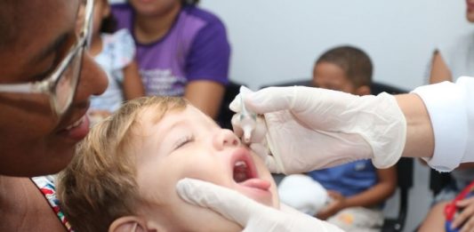 criança com os fechados e boca aberta pra cima coloca a língua pra fora ao receber gotas de vacina seguradas por mãos usando luvas; uma mulher segura a criança