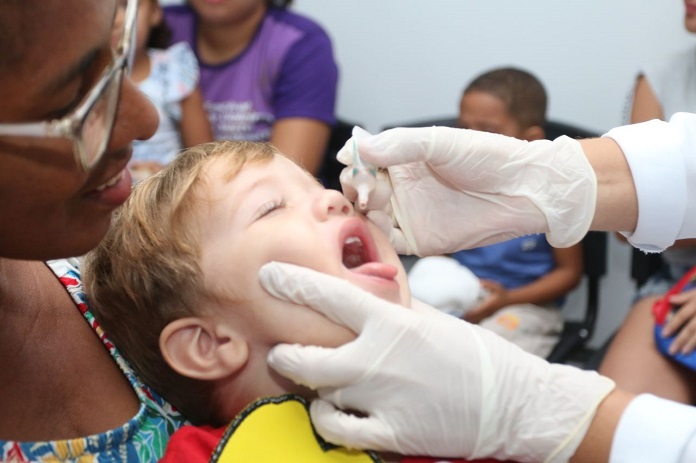 criança com os fechados e boca aberta pra cima coloca a língua pra fora ao receber gotas de vacina seguradas por mãos usando luvas; uma mulher segura a criança