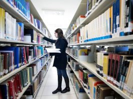 Conselho da Udesc aprova a criação de 12 cursos de pós-graduação mulher confere prateleira em um corredor no meio de estantes de livros em uma biblioteca