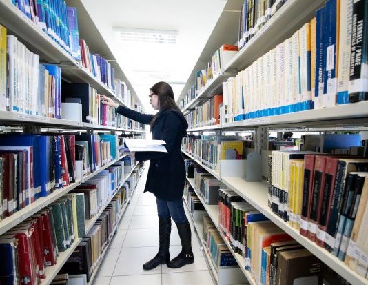 Conselho da Udesc aprova a criação de 12 cursos de pós-graduação mulher confere prateleira em um corredor no meio de estantes de livros em uma biblioteca
