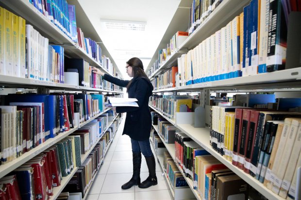 mulher confere prateleira em um corredor no meio de estantes de livros em uma biblioteca