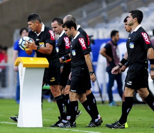 quatro árbitros com uniforme com propaganda na manga entram em campo juntos; o juiz pega a bola de cima de um pedestal no campo