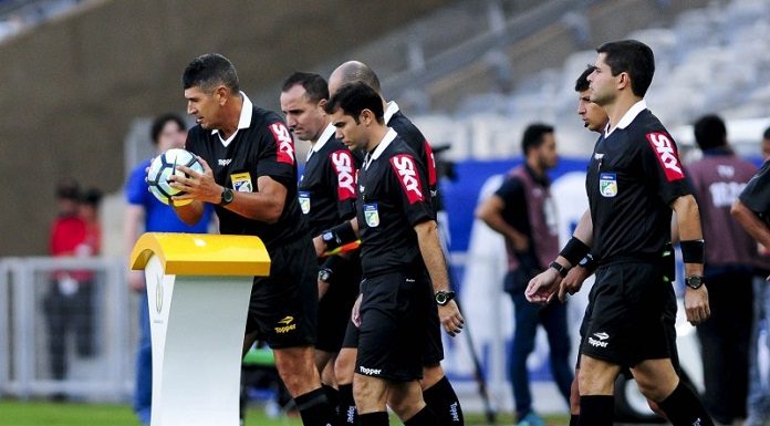 quatro árbitros com uniforme com propaganda na manga entram em campo juntos; o juiz pega a bola de cima de um pedestal no campo