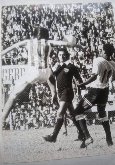 foto antiga de uma partida de futebol e preto e branco mostra árbitro acompanhando lance de cabeça com torcida ao fundo