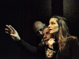 uma atriz e dois atores gesticulam olhando para o alto e um palco pouco iluminado