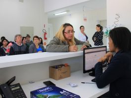 Inicia o funcionamento da UBS Prado, em Biguaçu mulher mostra a identidade sobre balcão para uma moça atendente sentada em frente à computador; outras pessoas sentadas esperam ao fundo
