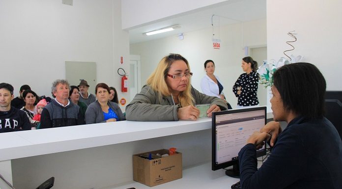 mulher mostra a identidade sobre balcão para uma moça atendente sentada em frente à computador; outras pessoas sentadas esperam ao fundo