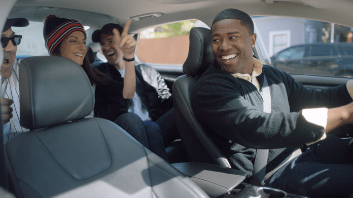 foto dentro de um carro; motorista abre largo sorrido olhando para 3 passageiros animados no banco de trás