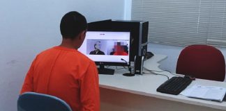 homem utilizando macacão laranja de detento é visto de costas sentado em frente a um computador com imagens de webcams de outras pessoas