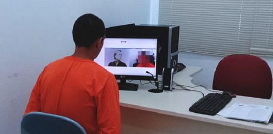 homem utilizando macacão laranja de detento é visto de costas sentado em frente a um computador com imagens de webcams de outras pessoas