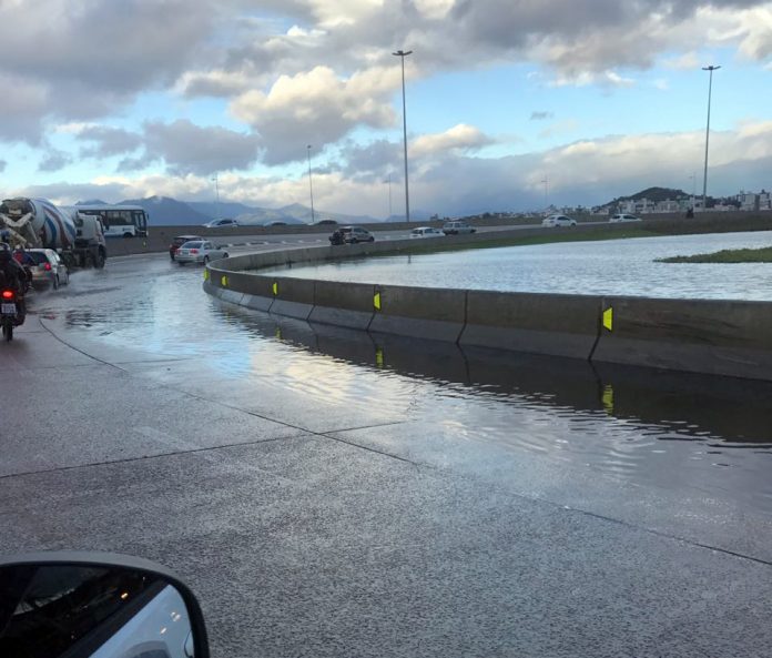 foto tirada de dentro de um carro mostra uma curva fechada, onde há um lago ao lado, com água na pista e alguns carros passando