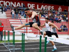 prova de atletismo masculino em que um homem salta a barreira e outros dois ao fundo se preparam também para pular; arquibancada mais ao fundo, onde se lê Lages