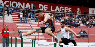 prova de atletismo masculino em que um homem salta a barreira e outros dois ao fundo se preparam também para pular; arquibancada mais ao fundo, onde se lê Lages