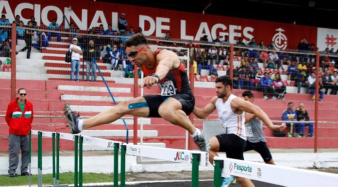 prova de atletismo masculino em que um homem salta a barreira e outros dois ao fundo se preparam também para pular; arquibancada mais ao fundo, onde se lê Lages