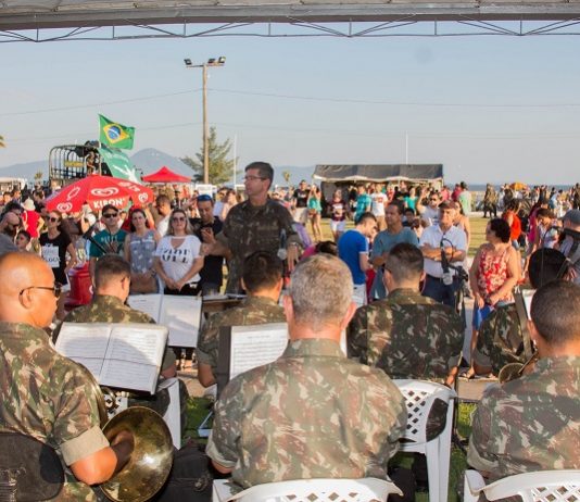 Cidades realizam desfile cívico neste domingo (2) homens com roupa camuflada do exército tocando em uma orquestra; maestro em pé de frente para câmera rege a música e populares ao fundo observam